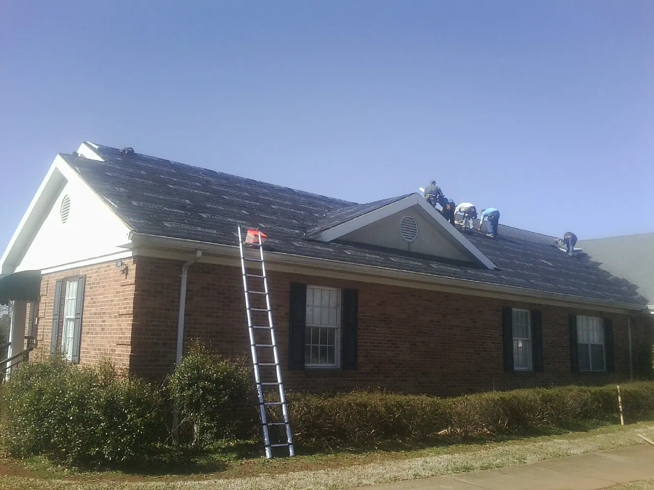 Crew tearing shingles off a brick ranch roof with a ladder set — Metal Roof Repair work by our crew in North Fork