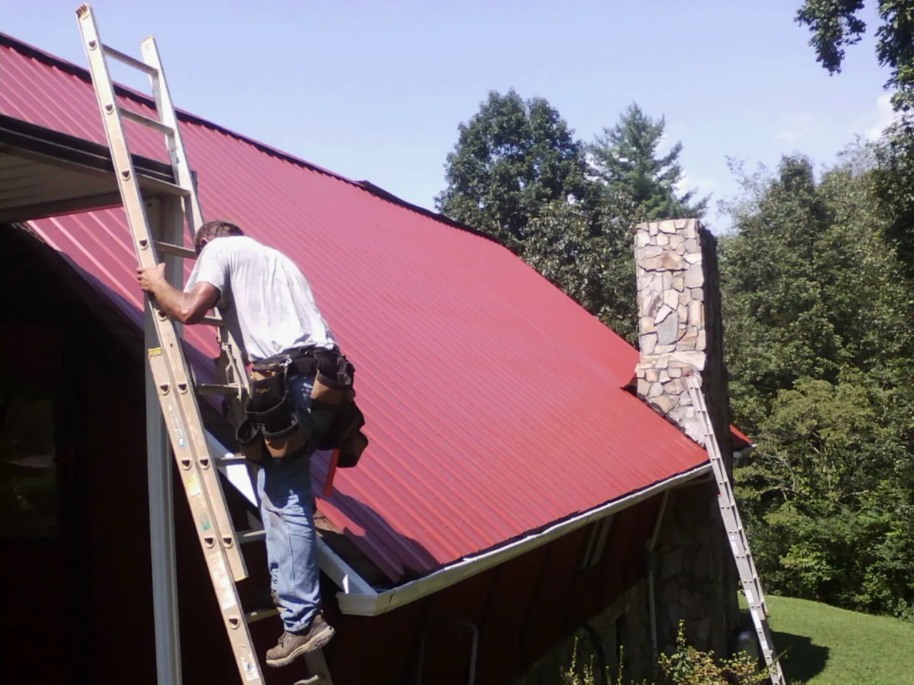 Roofer on a ladder beside a red metal roof and stone chimney — Soffit & Fascia Repair by Roofing Brothers in North Fork